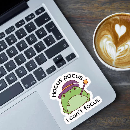 A die-cut sticker featuring a green cartoon frog wearing a purple hat and holding a wand, with the text 'Hocus pocus I can't focus', shown on a silver laptop next to a cup of latte on a dark wooden desk.