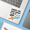 A die-cut sticker with the text 'CURRENT SIDE QUEST BROUGHT TO YOU BY ADHD' and a cartoon sword graphic, shown applied to the palm rest of a silver laptop on a light blue desk with a notebook nearby.