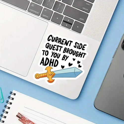 A die-cut sticker with the text 'CURRENT SIDE QUEST BROUGHT TO YOU BY ADHD' and a cartoon sword graphic, shown applied to the palm rest of a silver laptop on a light blue desk with a notebook nearby.
