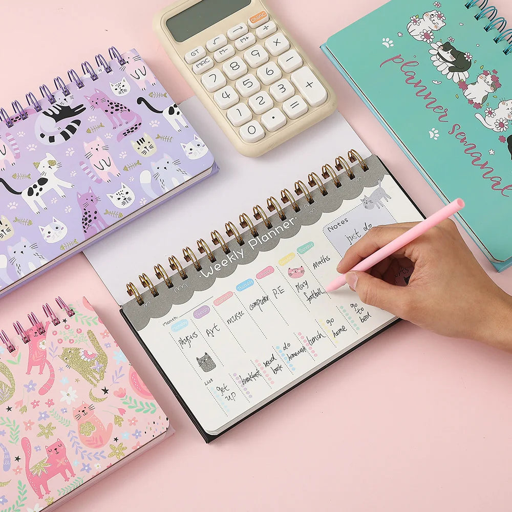 weekly planners with cute designs on a pink surface, one being used with a pen.